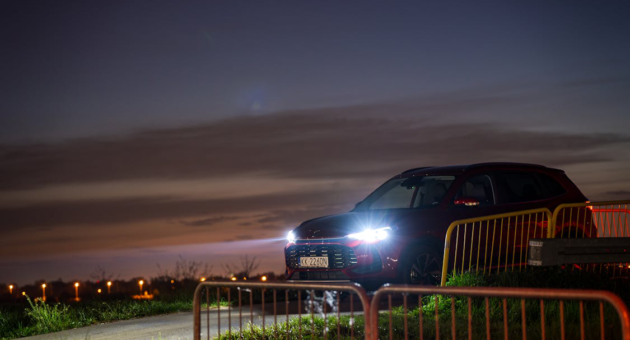 A red SUV with headlights on is parked along a rural road at sunset
