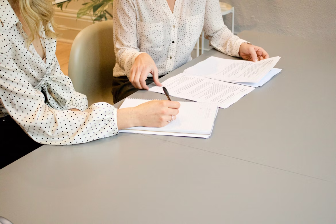 women-signing women wearing white shirts sitting at a table, going through documents, and signing