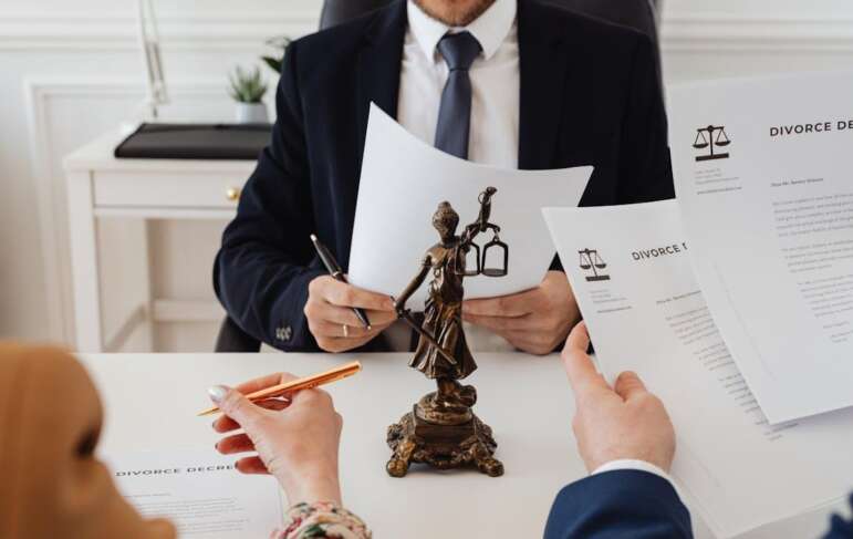 An image focused on a lady of justice statue placed in the middle of a table with three people having a meeting, holding divorce papers.
